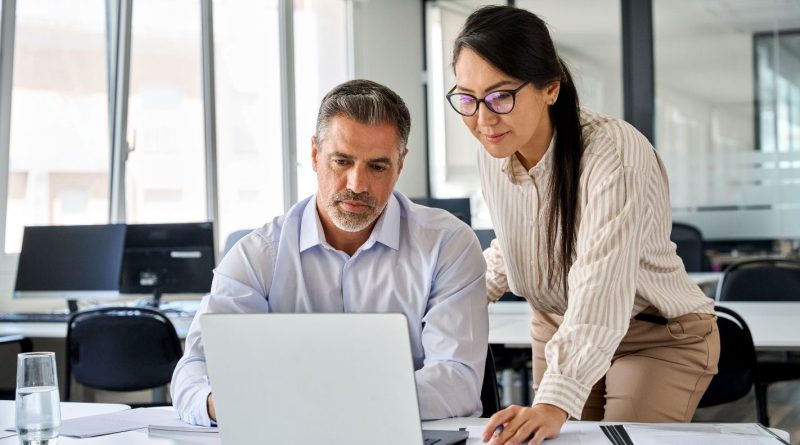 Deux collègues travaillent ensemble dans un bureau moderne : un homme assis regarde un ordinateur portable tandis qu’une femme debout à côté de lui consulte des documents.