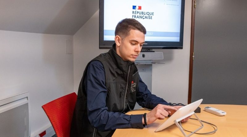 Un homme assis à une table utilise une tablette, avec un écran affichant « République française » en arrière-plan.