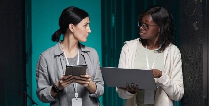Deux femmes marchent dans un couloir de serveurs informatiques et échangent en consultant une tablette et un ordinateur portable.