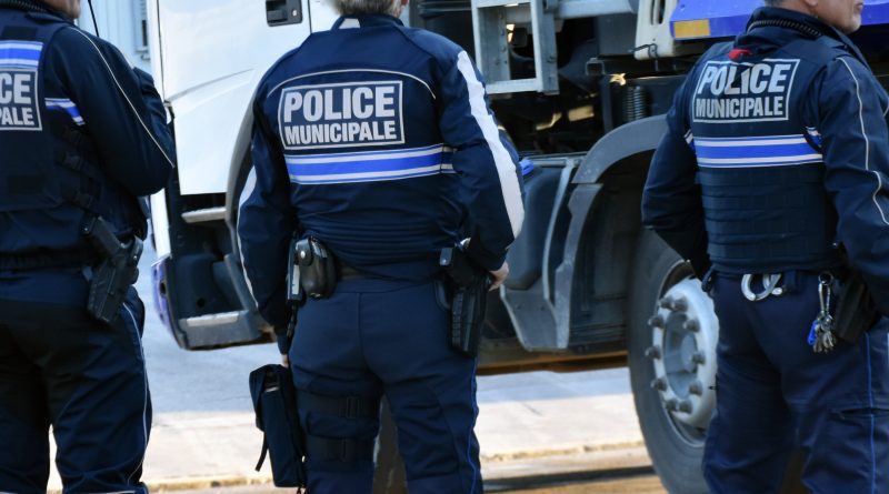 Trois agents de la police municipale vus de dos, en uniforme bleu, debout à proximité d’un véhicule de service. Leurs vestes portent l’inscription « Police municipale » et ils sont équipés de ceinturons avec matériel professionnel.