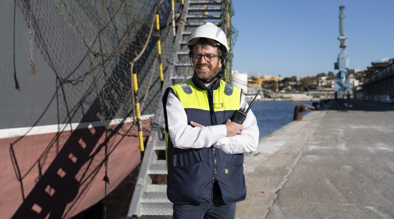 Homme portant un casque de sécurité et un gilet haute visibilité, tenant une radio, debout sur un quai de port à côté d’un navire, avec des installations portuaires en arrière-plan.