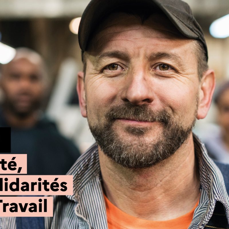 Portrait d’un homme portant une casquette et des vêtements de travail, regardant l’objectif, avec des collègues floutés en arrière-plan dans un environnement professionnel. Un message met en avant la santé, les solidarités et le travail.