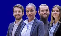 Quatre professionnels en tenue formelle, posant côte à côte devant un fond bleu uni, regardant l’objectif avec une expression confiante.