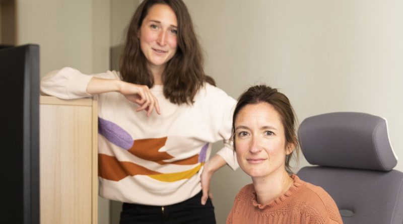 Deux femmes dans un bureau. L’une est assise à un bureau en train de travailler sur un ordinateur portable, tandis que l’autre se tient debout à côté d’elle. Un écran d’ordinateur, un clavier et des documents sont visibles sur le bureau.
