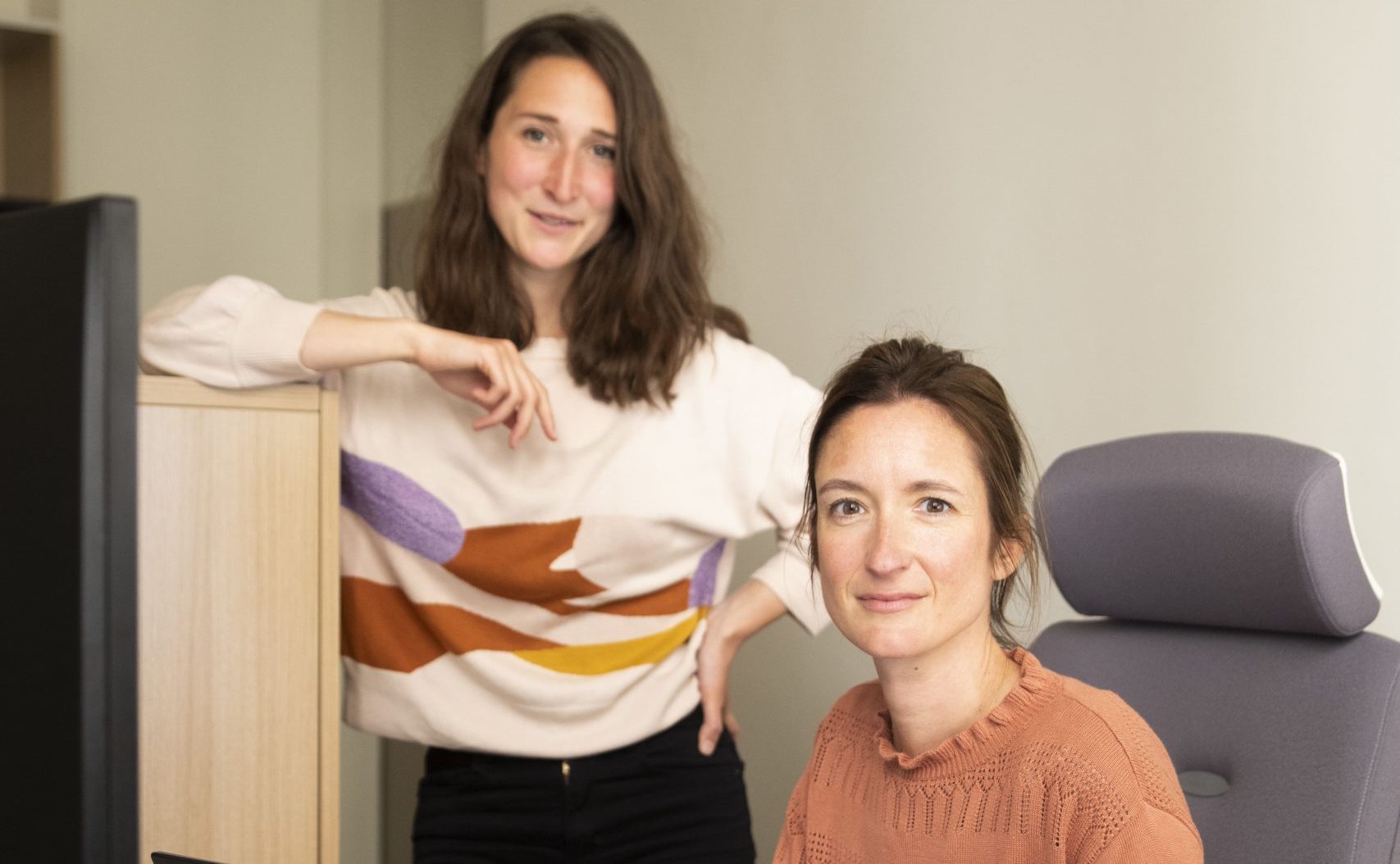 Deux femmes dans un bureau. L’une est assise à un bureau en train de travailler sur un ordinateur portable, tandis que l’autre se tient debout à côté d’elle. Un écran d’ordinateur, un clavier et des documents sont visibles sur le bureau.