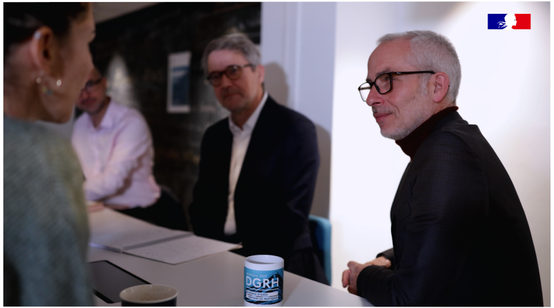 Un homme aux cheveux gris et lunettes est assis à une table lors d’une réunion professionnelle, en train d’échanger avec plusieurs personnes. Une tasse portant le logo « DGRH » est posée sur la table. La scène se déroule dans un espace de travail lumineux.