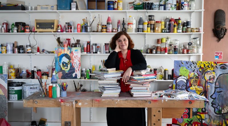 Au premier plan : Stéphanie, professeur technique culture et savoirs de base, dans son atelier. Elle s'appuie sur des livres posés sur un établi. Au second plan : des étagères sur lesquelles sont posés des pots de peintures et du matériel pour les arts plastiques. Un tableau est posé au sol.