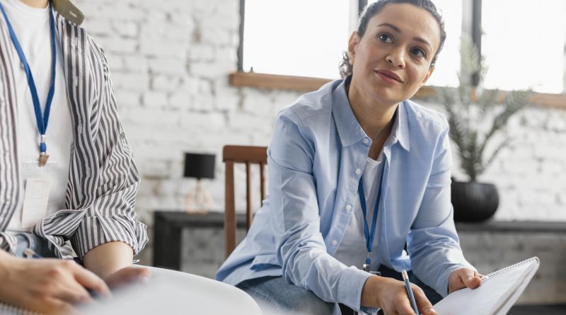 une femme assise tient un cahier. Elle écoute une personne dont on distingue la silhouette sur le bord gauche de la photo.
