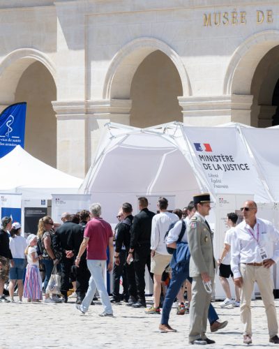 Une foule de visiteurs, dont des civils et des militaires en uniforme, se presse autour de du stand du ministère de la Justice et de l’administration pénitentiaire installé dans la cour d’honneur de l’Hôtel des Invalides à Paris en 2022. En arrière-plan, la façade du Musée de l’Armée avec ses arches et son inscription dorée.