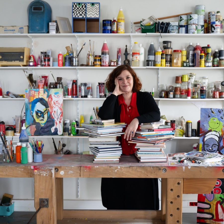 Au premier plan : Stéphanie, professeur technique culture et savoirs de base, dans son atelier. Elle s'appuie sur des livres posés sur un établi. Au second plan : des étagères sur lesquelles sont posés des pots de peintures et du matériel pour les arts plastiques. Un tableau est posé au sol.