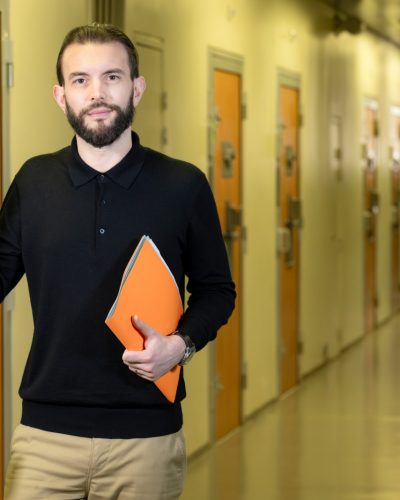 Au premier plan : un homme tenant dossier papier. Il ouvre une porte sur laquelle est écrit bureau d’audience. Au second plan, un couloir de prison, les portes des cellules sont fermées. Au fond, on distingue un surveillant pénitentiaire vérifiant une porte de cellule.