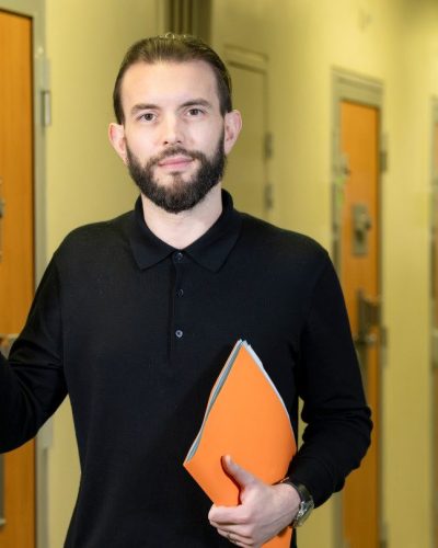 Au premier plan : un homme tenant dossier papier. Il ouvre une porte sur laquelle est écrit bureau d’audience. Au second plan, un couloir de prison, les portes des cellules sont fermées. Au fond, on distingue un surveillant pénitentiaire vérifiant une porte de cellule.
